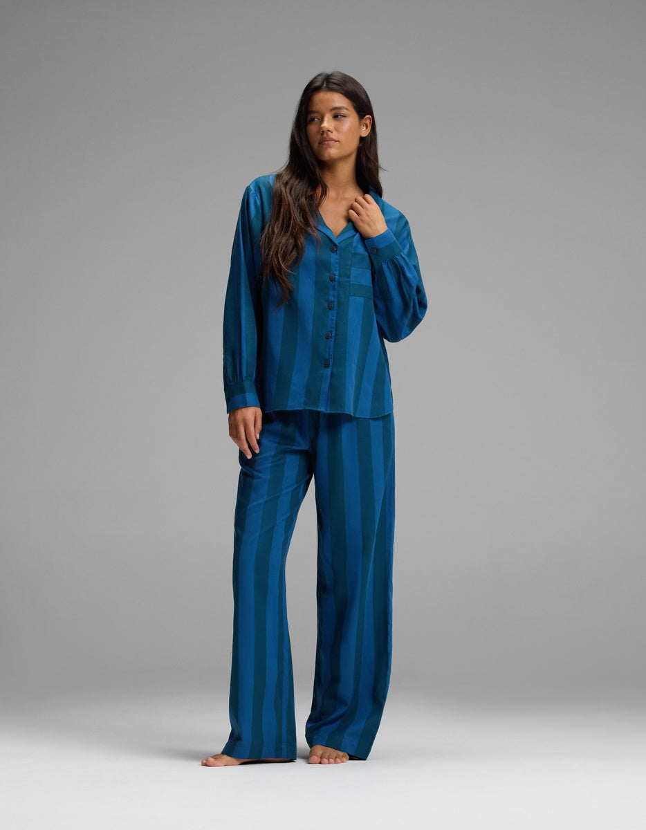 A woman stands barefoot against a plain gray background, wearing the Cozy Earth Women’s Belmont Long Sleeve Pajama Top paired with matching loose-fitting blue striped pajama pants.