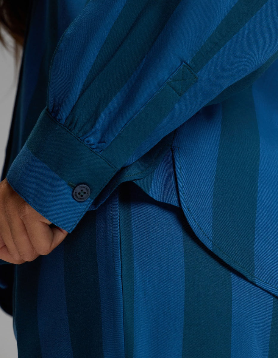 Close-up of a person wearing the Cozy Earth Women’s Belmont Long Sleeve Pajama Top, featuring blue fabric with dark blue vertical stripes and matching pants. The shirt cuff is buttoned, and the person’s hand is visible near the side seam.