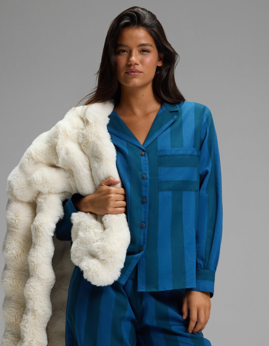 A woman wearing the Cozy Earth Women’s Belmont Long Sleeve Pajama Top drapes a white faux fur blanket over her shoulder, standing against a plain gray background.
