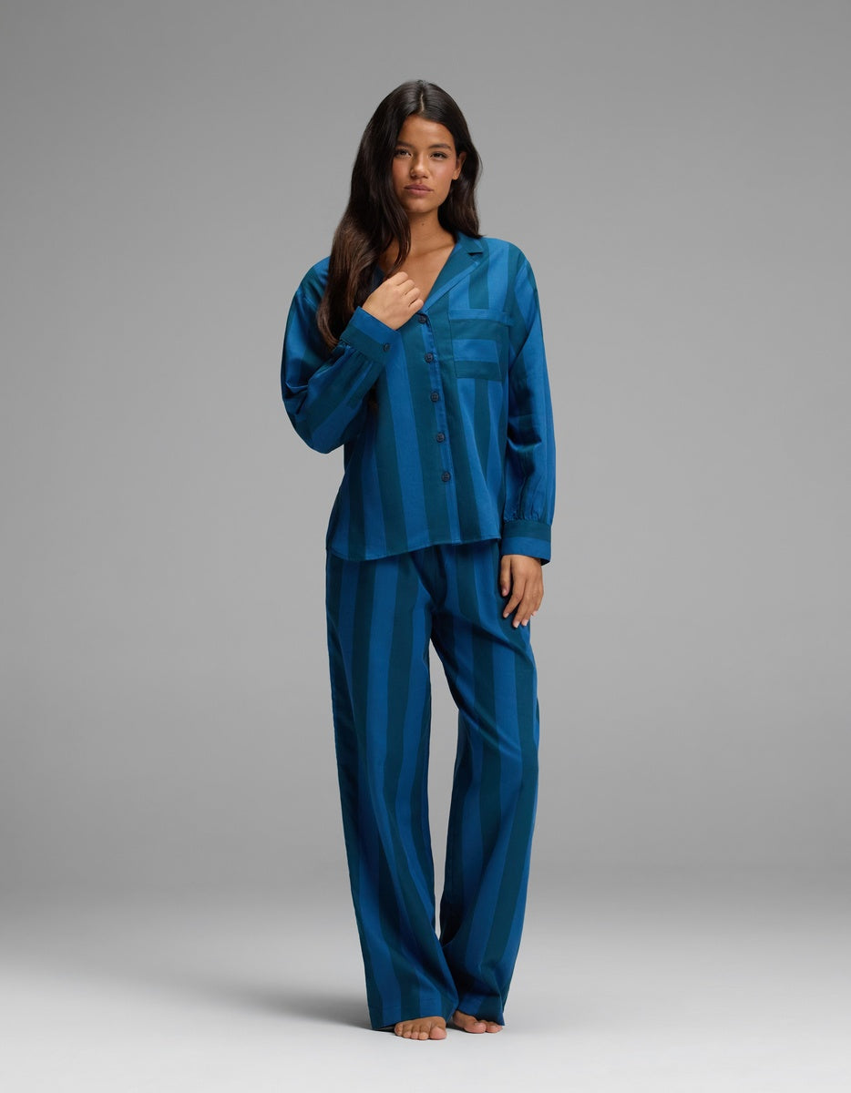 A woman stands barefoot against a plain gray background, wearing the Cozy Earth Women’s Belmont Long Sleeve Pajama Set in Anchor Stripe.