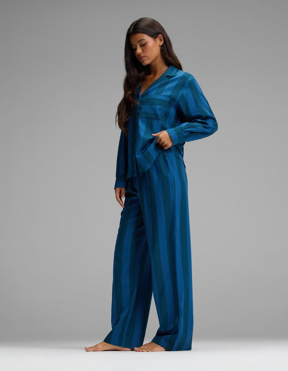 A woman stands barefoot against a plain gray background, wearing the Cozy Earth Women’s Belmont Long Sleeve Pajama Set—loose-fitting, blue striped pajamas featuring wide-leg pants and long sleeves, with one hand in her pocket.