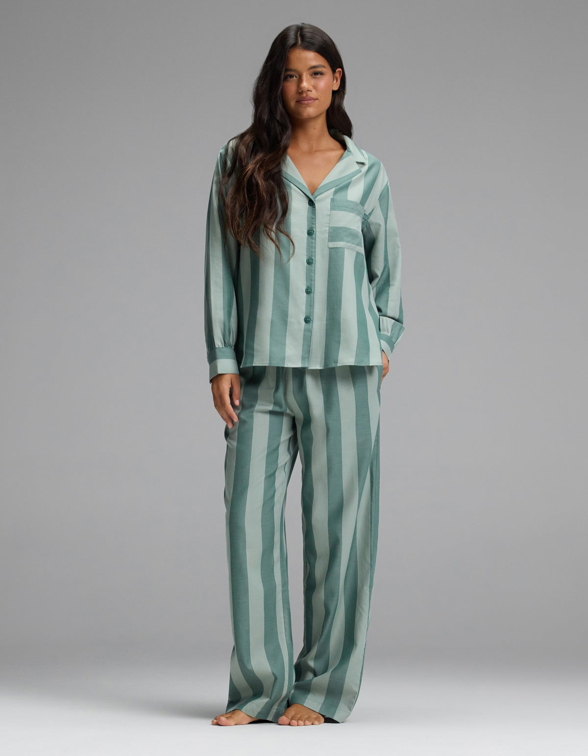 A woman stands barefoot against a plain gray background, wearing the Cozy Earth Women’s Belmont Long Sleeve Pajama Set in Rosemary Stripe—featuring loose-fitting, vertically striped green and light green button-up top with matching pants.|Color:Rosemary Stripe