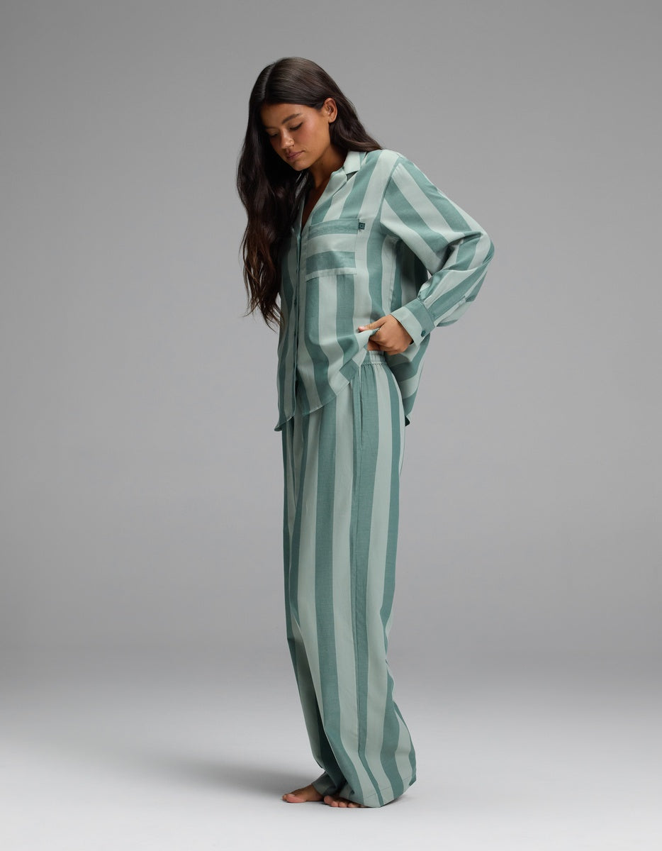 A woman models the Cozy Earth Women’s Belmont Long Sleeve Pajama Set in green and white stripes, standing barefoot against a plain gray background with her hand in her pocket, gazing downward.