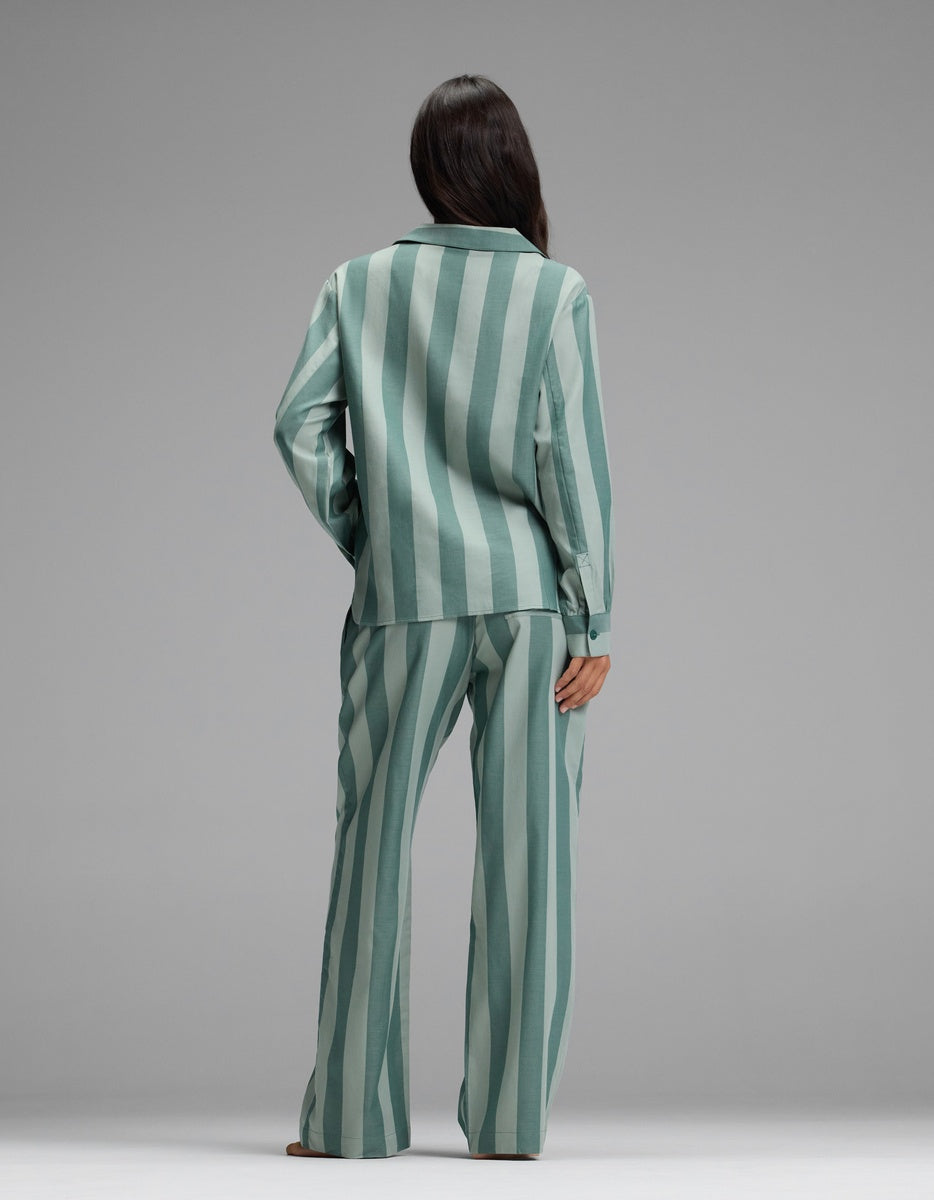 A woman stands facing away, wearing the Cozy Earth Women’s Belmont Long Sleeve Pajama Set in loose-fitting, green and light teal vertical stripes. The background is plain gray.