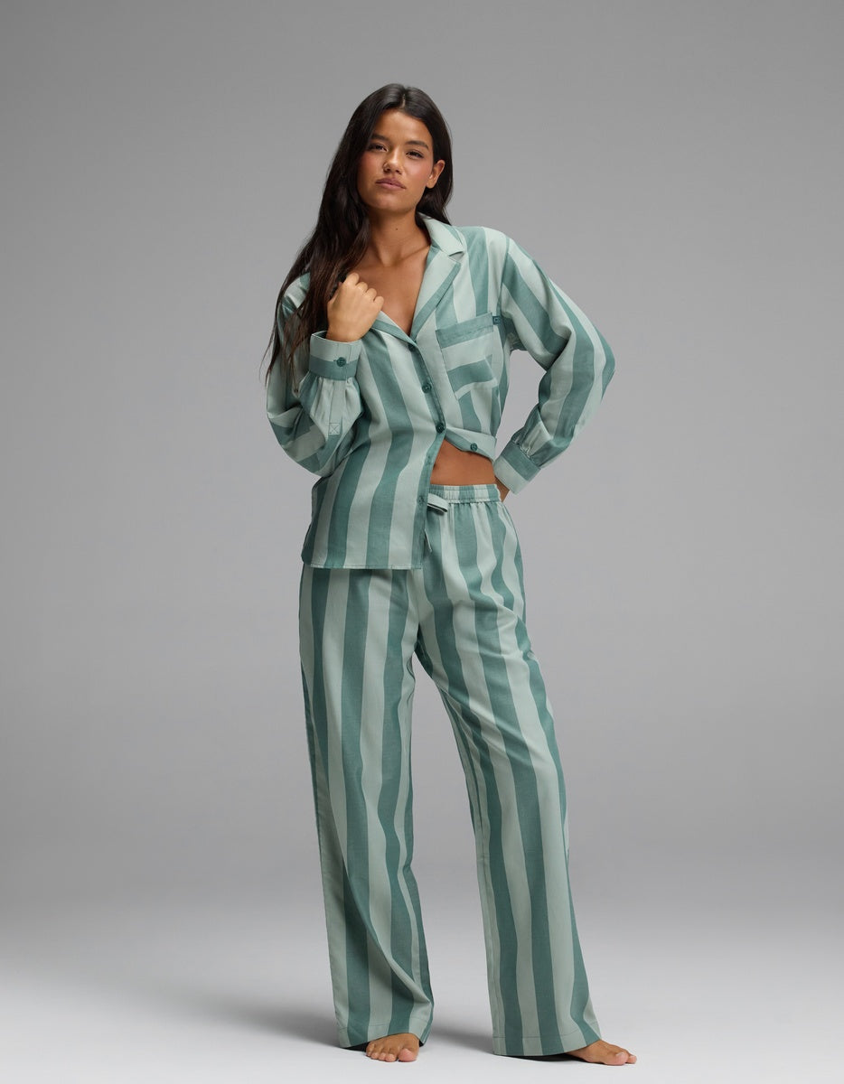 A woman stands barefoot against a plain grey background, posing with one hand on her hip and modeling the Cozy Earth Women’s Belmont Long Sleeve Pajama Set in a loose, green and teal vertically striped design with long sleeves and matching pants.