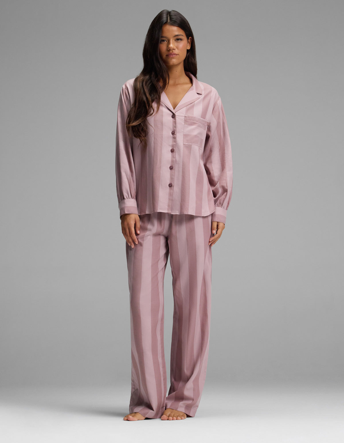 A woman stands barefoot, facing forward against a plain gray background, wearing the Cozy Earth Women’s Belmont Long Sleeve Pajama Set in Viola Stripe. The loose-fitting, button-up pajamas feature long sleeves and matching pants with a vertical stripe pattern.