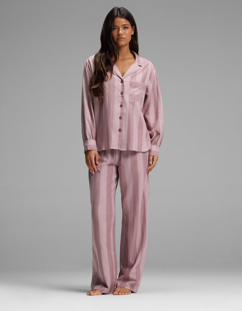 A woman stands barefoot, facing forward against a plain gray background, wearing the Cozy Earth Women’s Belmont Long Sleeve Pajama Set in Viola Stripe. The loose-fitting, button-up pajamas feature long sleeves and matching pants with a vertical stripe pattern.