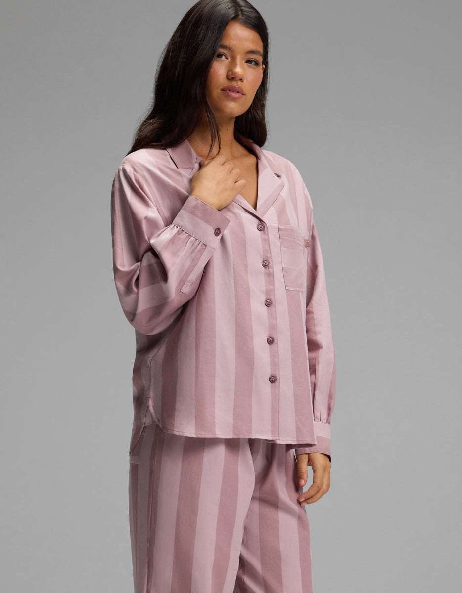 A person stands against a plain gray background wearing the Cozy Earth Women’s Belmont Long Sleeve Pajama Set, featuring a loose-fitting, button-up top and matching striped pants in light mauve.
