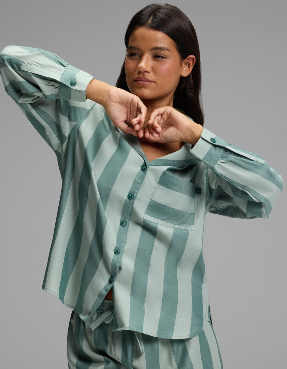A woman wearing the Cozy Earth Women’s Belmont Long Sleeve Pajama Top in a loose, green and light green vertically striped pattern, along with matching pants, poses with her hands raised near her face against a plain gray background.|Color:Rosemary Stripe
