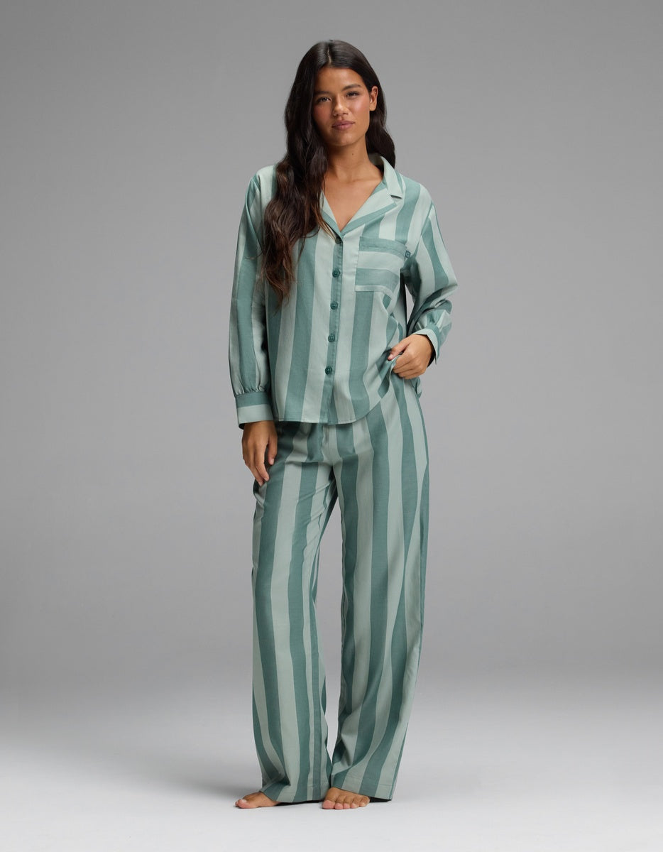 A woman stands barefoot against a plain, light gray background, wearing the Cozy Earth Women’s Belmont Long Sleeve Pajama Top with matching loose green striped pajama pants. She has long dark hair and a neutral expression.