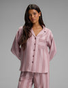 A woman with long dark hair wears the Cozy Earth Women’s Belmont Long Sleeve Pajama Top—a matching pink and mauve striped button-up pajama top with a chest pocket—while standing against a plain gray background.|Color:Viola Stripe