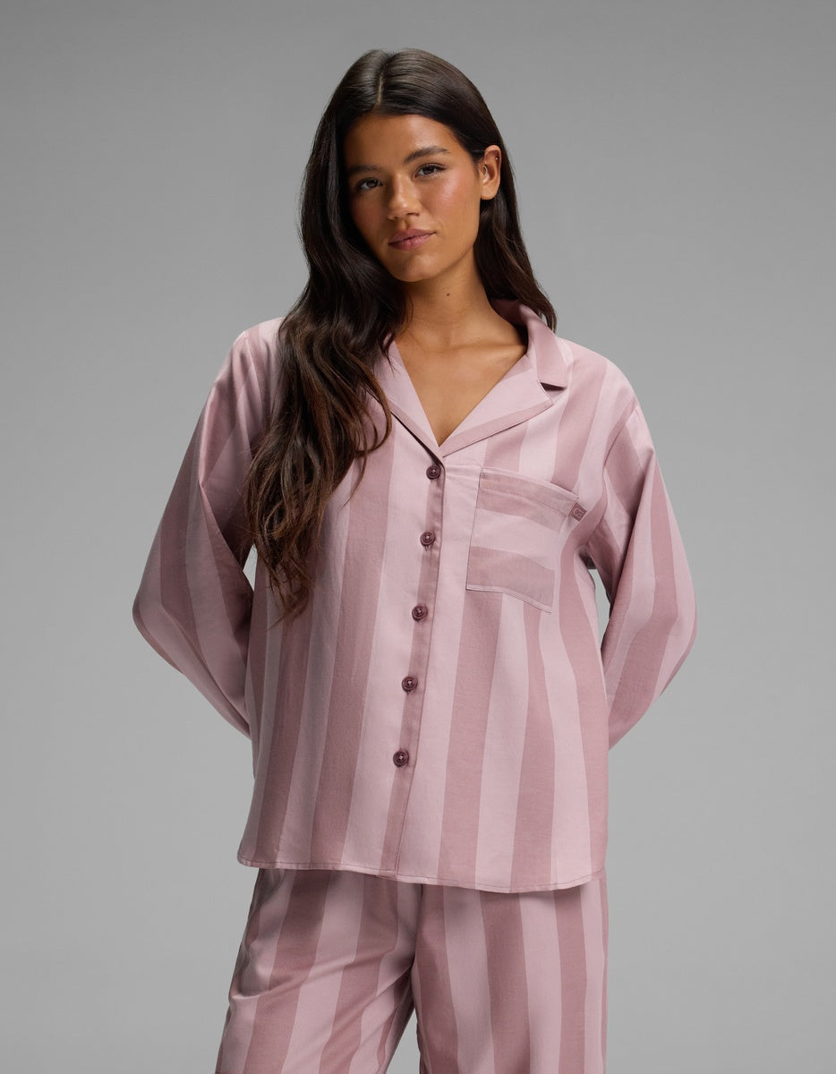 A woman with long dark hair wears the Cozy Earth Women’s Belmont Long Sleeve Pajama Top—a matching pink and mauve striped button-up pajama top with a chest pocket—while standing against a plain gray background.