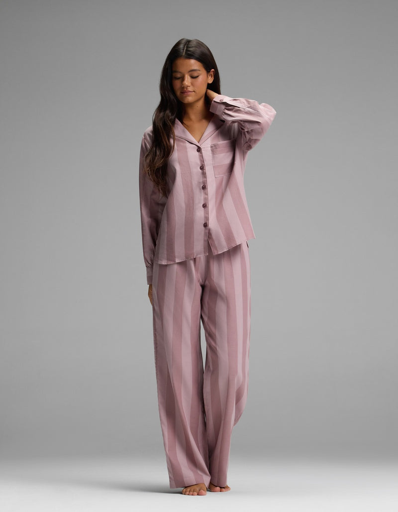 A woman stands barefoot on a gray background, wearing the Cozy Earth Women’s Belmont Long Sleeve Pajama Top in a loose mauve stripe pattern, paired with matching long pants. She has long dark hair and gently touches her neck with one hand.