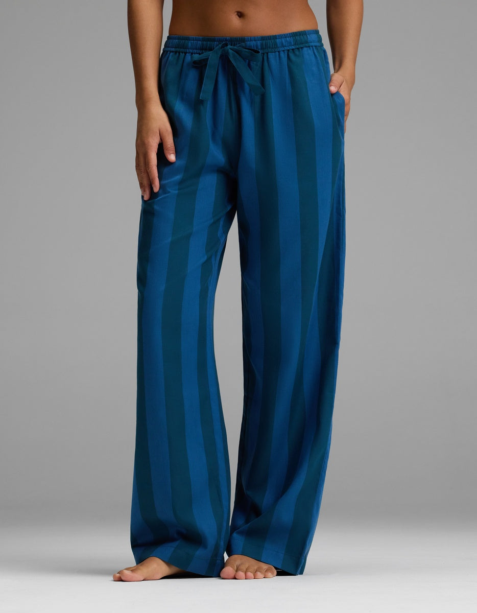 A person stands barefoot against a plain gray background wearing Cozy Earth’s Women’s Belmont Pajama Pant—a loose-fitting, blue striped style with a comfortable drawstring waist and hands relaxed by their sides.|Color:Anchor Stripe