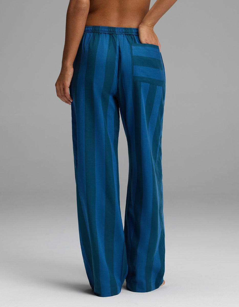A woman is shown from the back wearing Cozy Earth’s Women’s Belmont Pajama Pant in a loose-fitting blue striped design, featuring an elastic waistband and a single back pocket, with one hand resting on her hip.