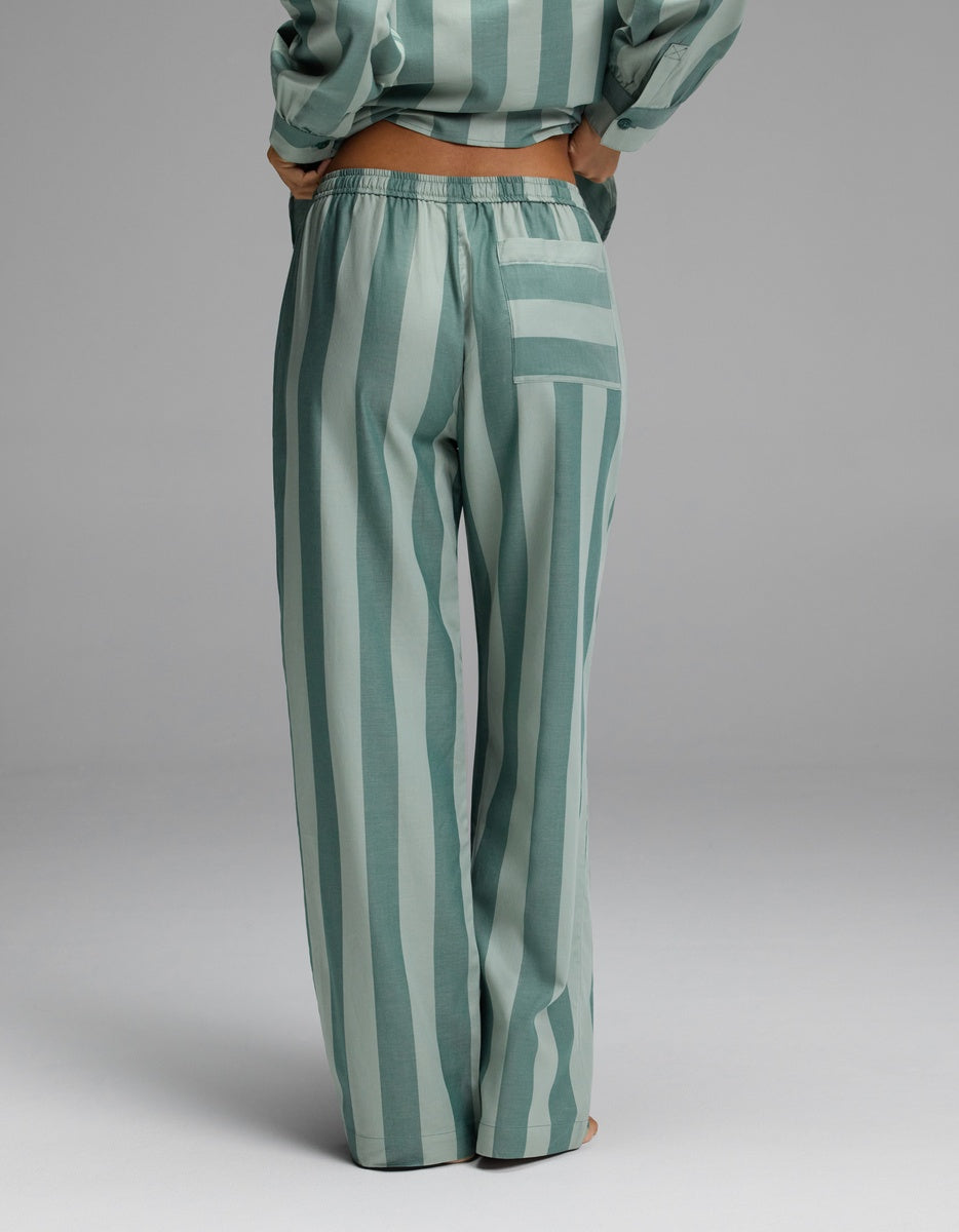 A person stands with their back to the camera, wearing the Cozy Earth Women’s Belmont Pajama Pant—a loose, high-waisted style in green and light green stripes—paired with a matching top. The pants feature a right-side pocket with horizontal stripes.|Color:Rosemary Stripe