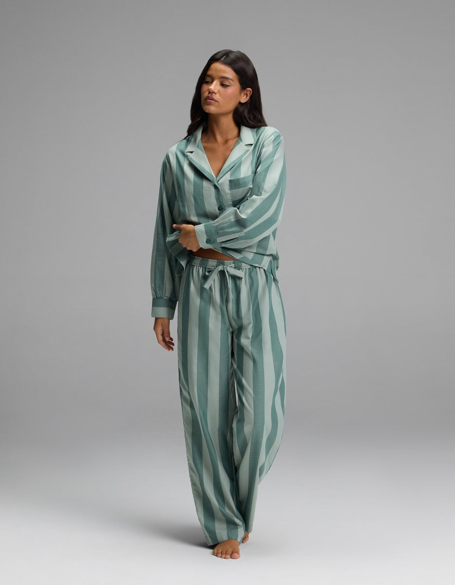 A woman with long dark hair stands barefoot against a plain gray background, gently crossing her arms in front of her body while wearing Cozy Earth’s Women’s Belmont Pajama Pant in a loose, green-and-white vertical stripe pattern.
