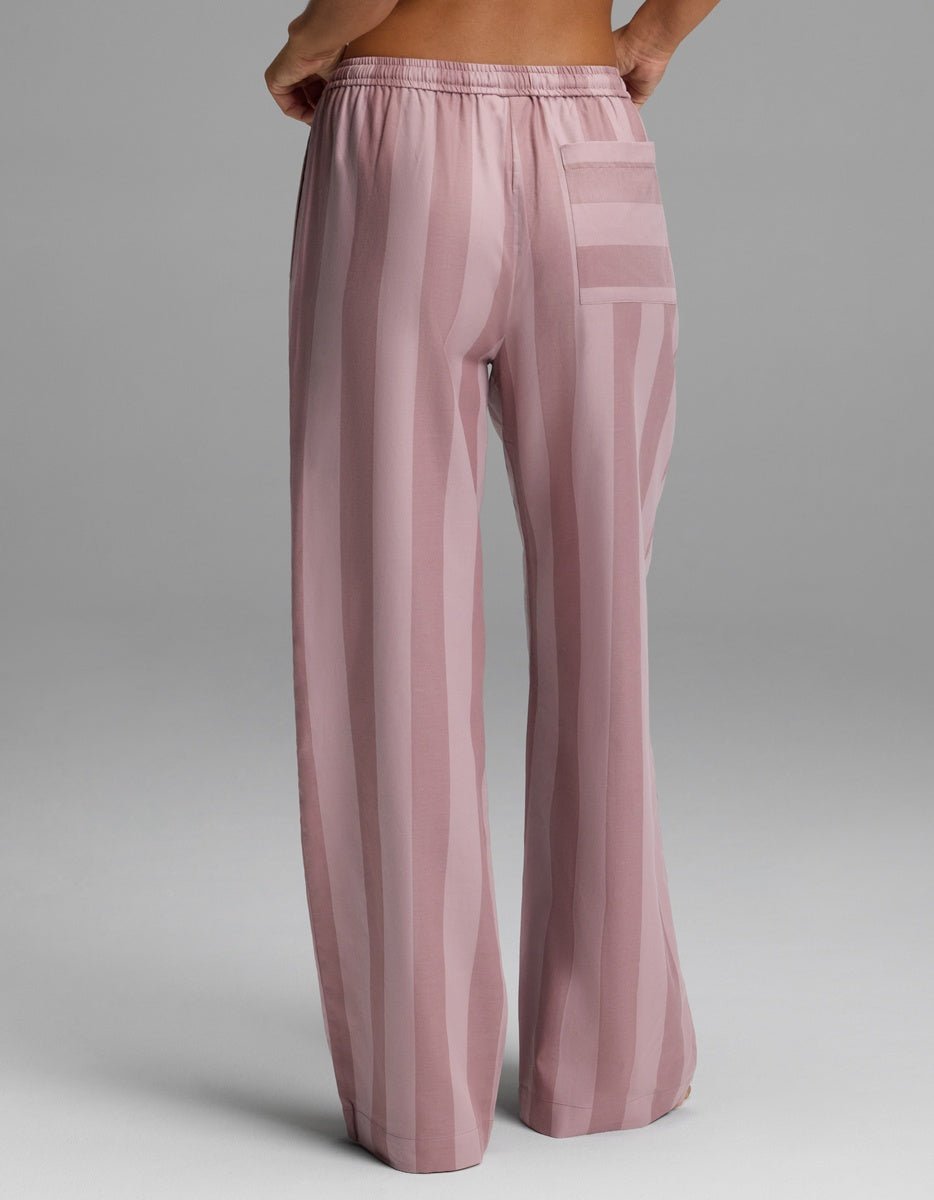 A woman is shown from behind against a plain gray background, wearing Cozy Earth’s Women’s Belmont Pajama Pant—loose, high-waisted pink pants with vertical stripes and a back pocket.