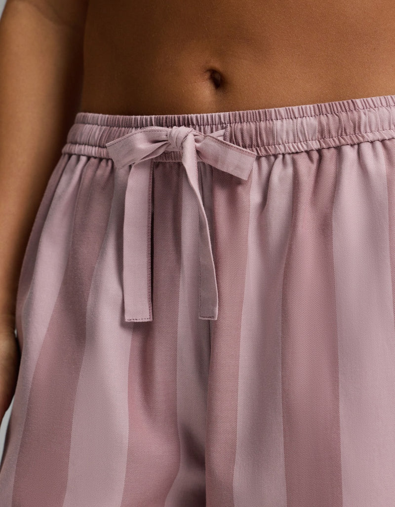 Close-up of a person’s midsection wearing Cozy Earth’s Women’s Belmont Pajama Pant, featuring silky, loose, light mauve stripes and a drawstring bow at the waistband. The person’s bare skin and belly button are visible above the pants.