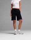 A person stands against a plain grey background wearing Cozy Earth Women’s Brushed Bamboo Bermuda Shorts, paired with a white fitted t-shirt, white socks, and white sneakers. Their hands are in their pockets and their face is not visible.|Color:Black