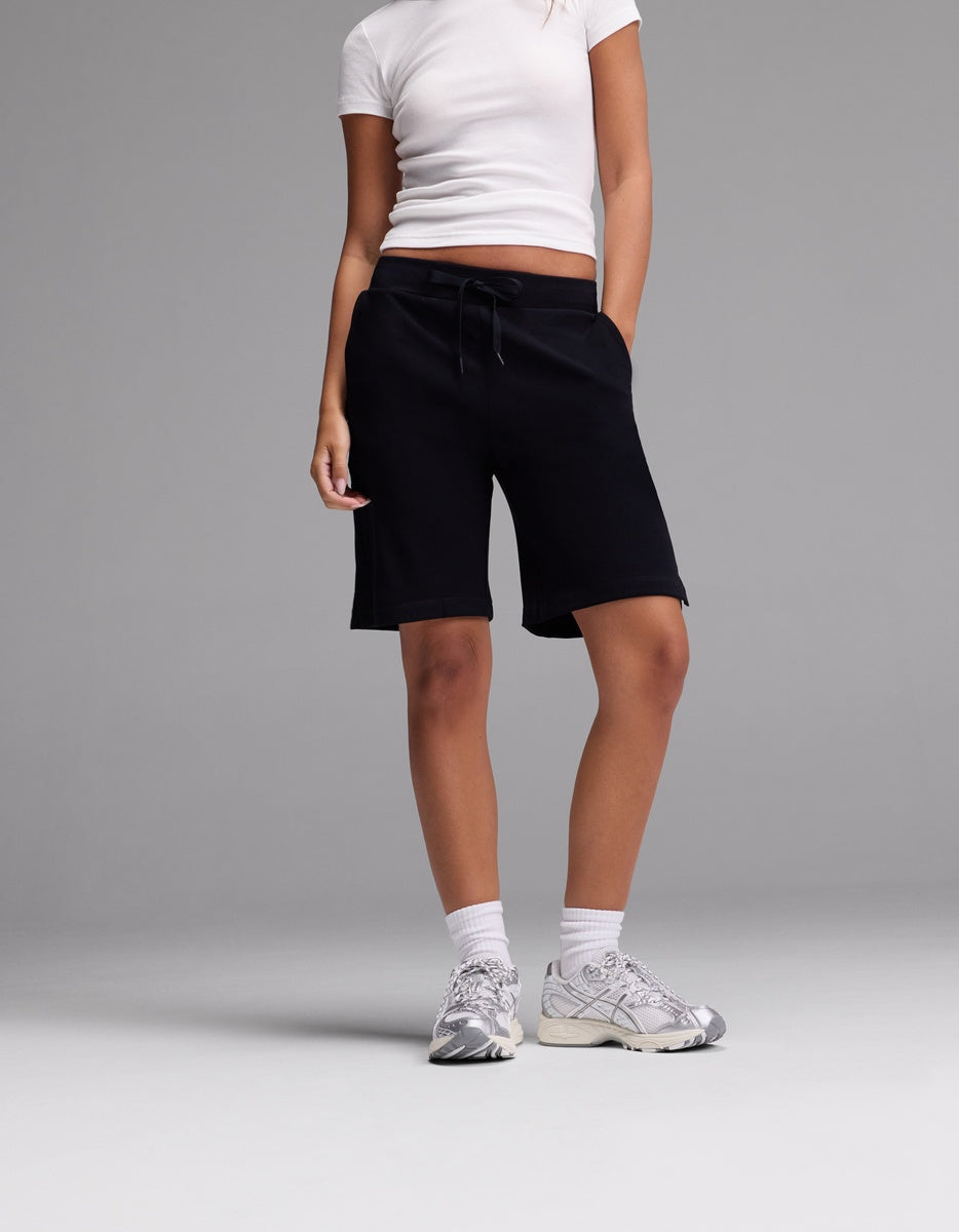 A person stands against a plain grey background wearing Cozy Earth Women’s Brushed Bamboo Bermuda Shorts, paired with a white fitted t-shirt, white socks, and white sneakers. Their hands are in their pockets and their face is not visible.|Color:Black