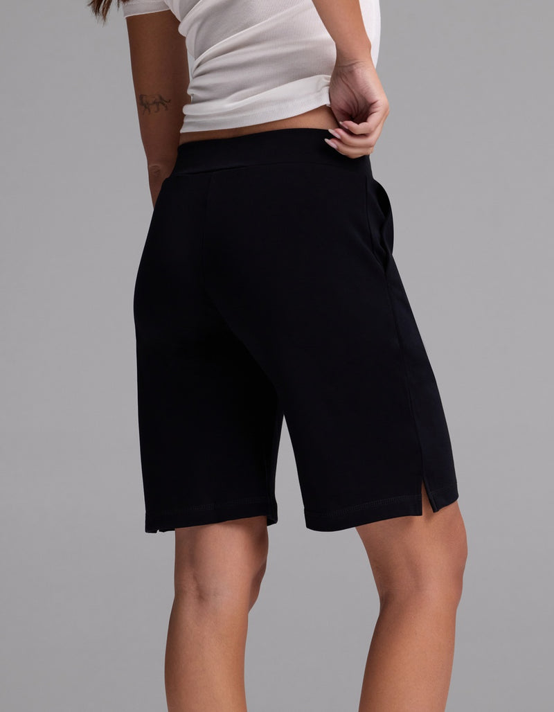 A person wearing a white shirt and Cozy Earth Women’s Brushed Bamboo Bermuda Shorts in black stands with one hand on their hip, facing away from the camera against a plain gray background.