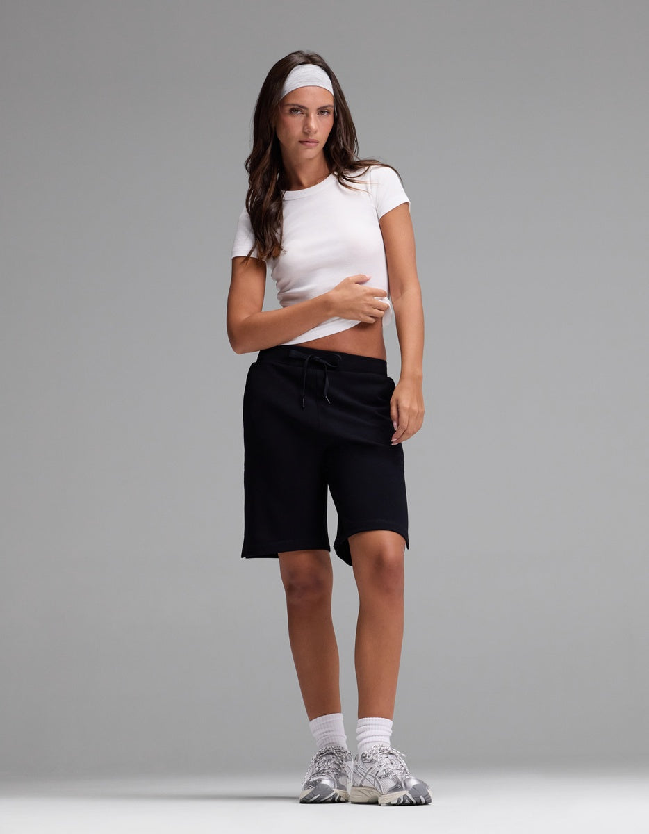 A person stands against a plain gray background wearing Cozy Earth’s Women’s Brushed Bamboo Bermuda Shorts, paired with a white headband, white crop top, white socks, and gray sneakers.