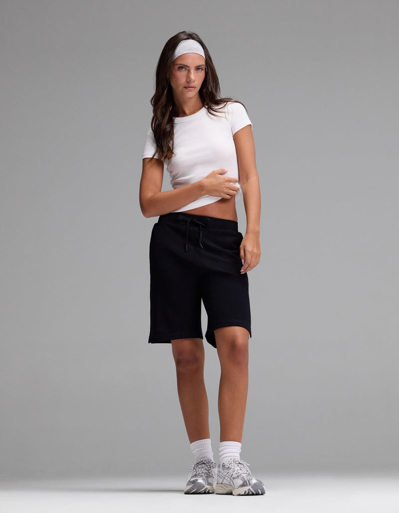 A person stands against a plain gray background wearing Cozy Earth’s Women’s Brushed Bamboo Bermuda Shorts, paired with a white headband, white crop top, white socks, and gray sneakers.
