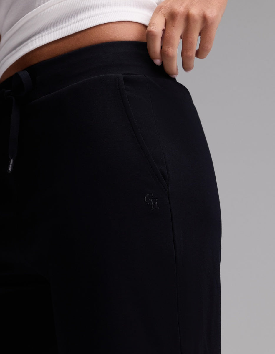 A person wearing Cozy Earth’s Women’s Brushed Bamboo Bermuda Shorts in black and a white top, with one hand resting on their hip. The shorts feature an embroidered “G B” logo near the pocket.