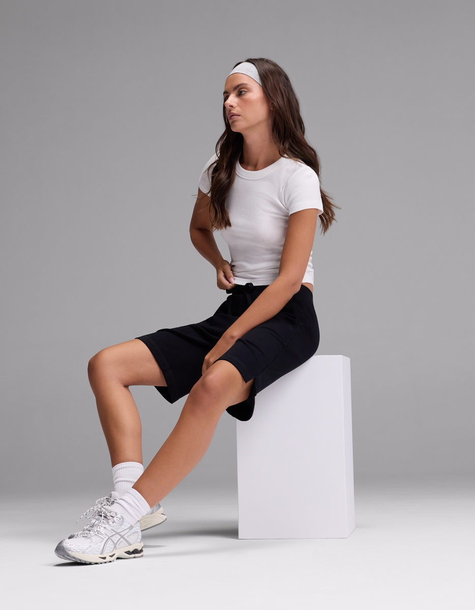 A woman wearing Cozy Earth’s Women’s Brushed Bamboo Bermuda Shorts, paired with a white t-shirt, white sneakers, and a gray headband, sits on a white block against a plain gray background, looking to the side.