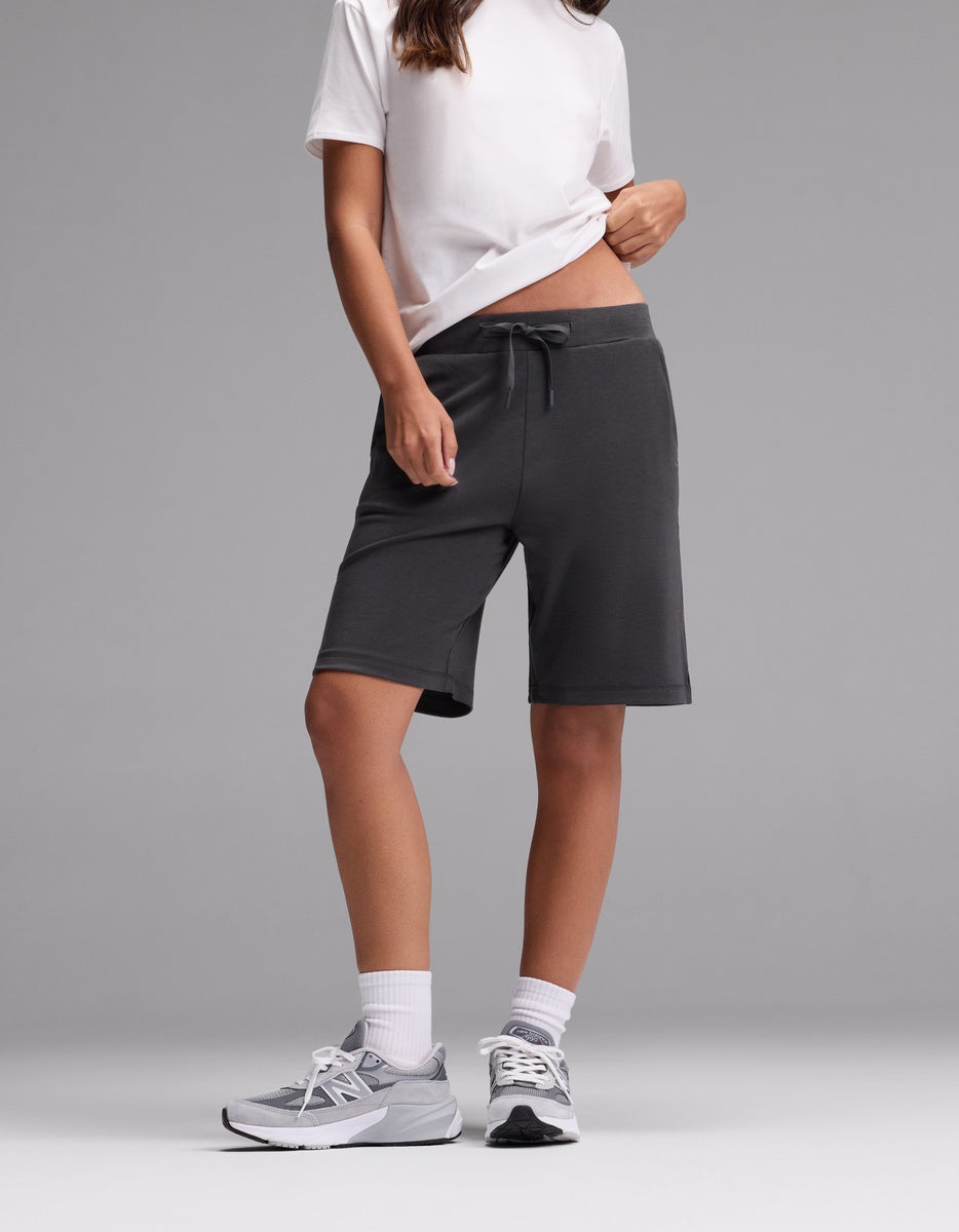 A person wearing a white t-shirt, Cozy Earth Women’s Brushed Bamboo Bermuda Shorts in dark gray, white socks, and gray sneakers stands against a plain gray background. Their face is not visible.