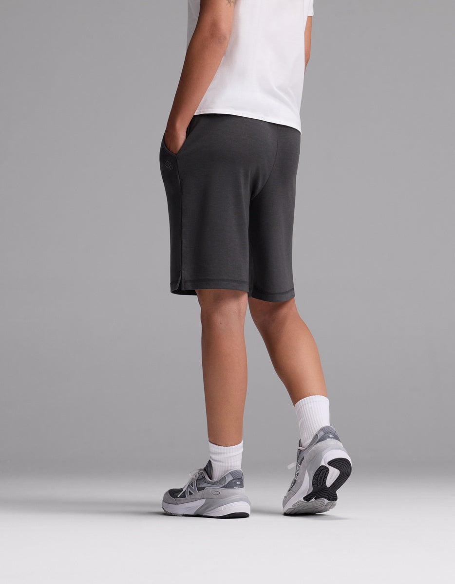 A person models Cozy Earth’s Women’s Brushed Bamboo Bermuda Shorts in gray, styled with a white t-shirt, white socks, and gray sneakers. They stand facing away against a plain gray background with one hand in their pocket.