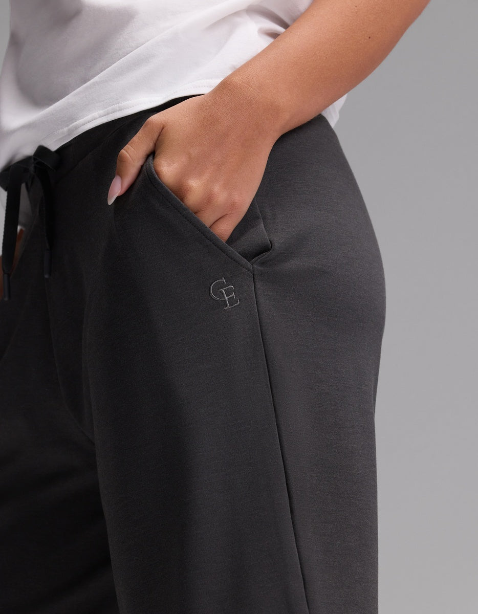 A person is wearing Cozy Earth’s Women’s Brushed Bamboo Bermuda Shorts in dark gray, paired with a white shirt and one hand in their pocket. The shorts feature a small embroidered logo near the pocket. The background is plain and gray.