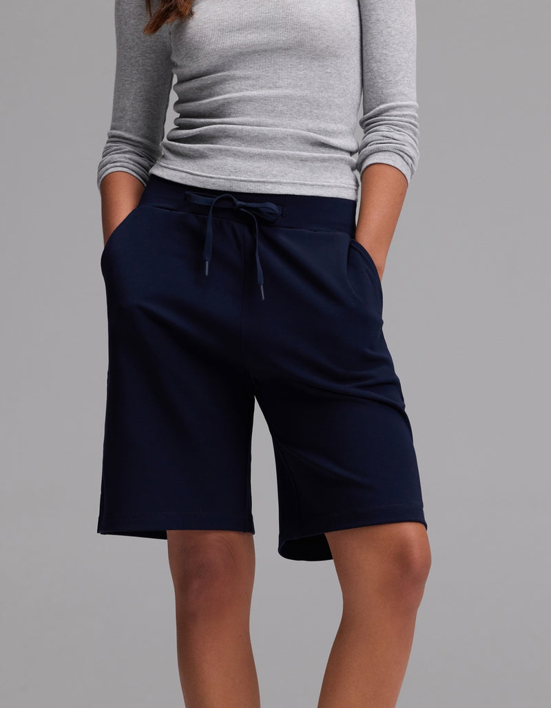 A person wearing a light gray long-sleeve shirt and Cozy Earth’s Women’s Brushed Bamboo Bermuda Shorts in dark navy, with hands in pockets, standing against a plain gray background.
