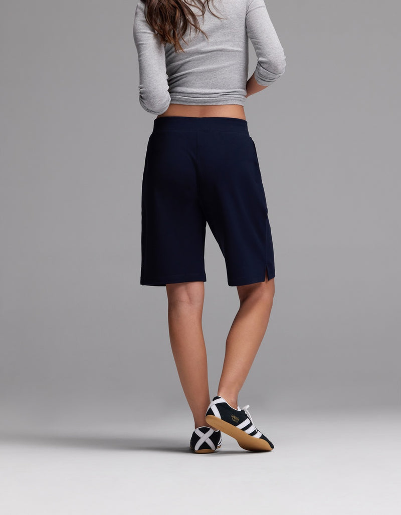 A person stands with their back to the camera, wearing a gray long-sleeve top, Cozy Earth Women’s Brushed Bamboo Bermuda Shorts in navy, and black-and-white sneakers against a plain gray background.