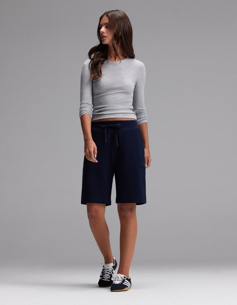 A woman stands against a plain gray background wearing a fitted gray long-sleeve top, black sneakers, and Cozy Earth’s Women’s Brushed Bamboo Bermuda Shorts in navy with a drawstring waist.