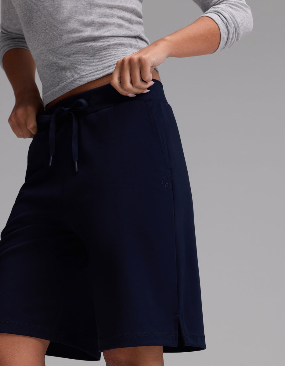 A person is shown from the shoulders down, set against a plain gray background, wearing Cozy Earth’s Women’s Brushed Bamboo Bermuda Shorts in navy blue with a drawstring waistband, paired with a light gray long-sleeve top; one hand is adjusting the shorts’ waistband.