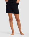 A person stands barefoot on a light gray surface, wearing Cozy Earth's Women's Brushed Bamboo Bermuda Shorts in black. The image is cropped from the waist down, highlighting the shorts and legs.|Color:Black
