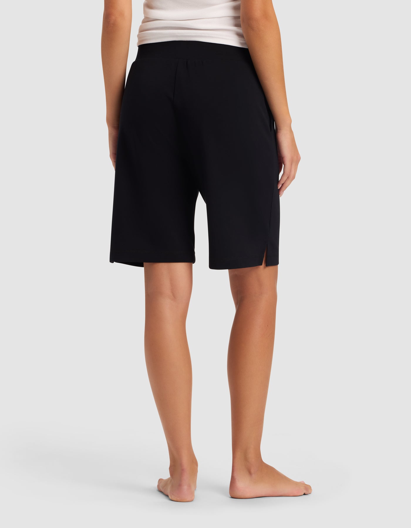 A woman stands barefoot on a white floor, seen from the back, wearing a white tank top and Cozy Earth Women's Brushed Bamboo Bermuda Shorts in black with a small side slit.