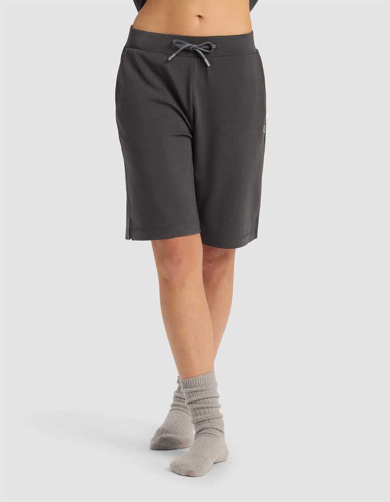 A person wearing Cozy Earth Women's Brushed Bamboo Bermuda Shorts in dark gray and light gray socks stands with legs crossed against a plain light background. The top half of their body is not visible.