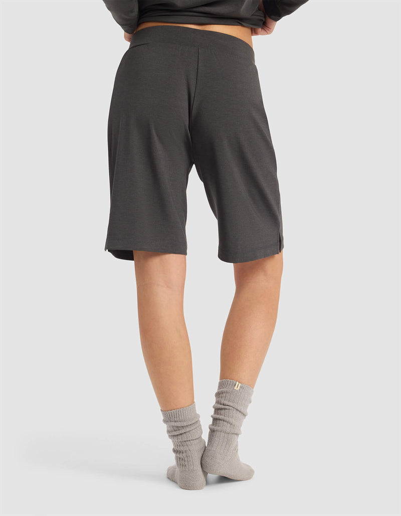 A woman, seen from behind on a white surface, wears Cozy Earth's Women's Brushed Bamboo Bermuda Shorts in dark gray. Only her lower body and part of her torso are visible, paired with light gray socks and a dark top.