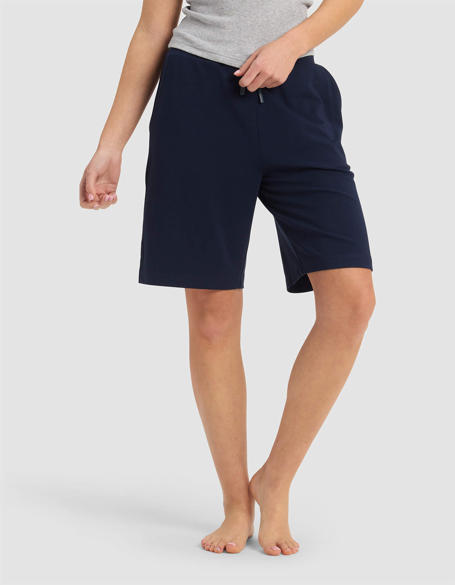 A person stands barefoot on a plain white background, wearing Cozy Earth’s Women’s Brushed Bamboo Bermuda Shorts in navy. The upper body is partially out of frame.