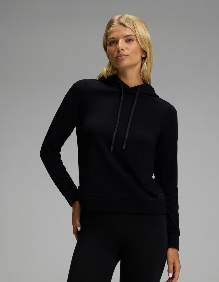 A woman with long blonde hair wears the Cozy Earth Women’s Brushed Bamboo Hoodie in black, paired with black pants, standing against a plain gray background with a neutral expression.|Color:Black