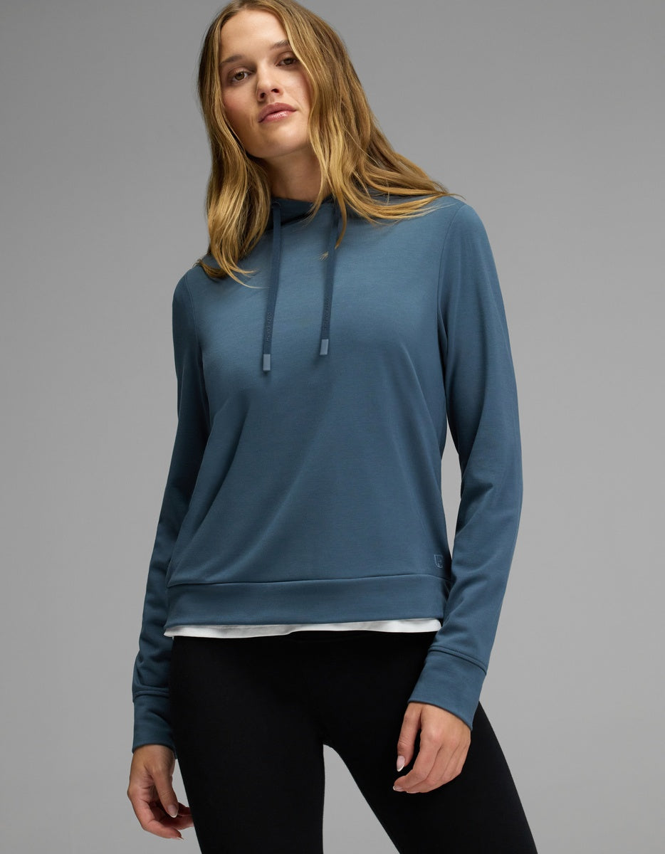 A woman with long, light brown hair wears the Cozy Earth Women’s Brushed Bamboo Hoodie in blue and black leggings, standing against a plain gray background.|Color:Dawn