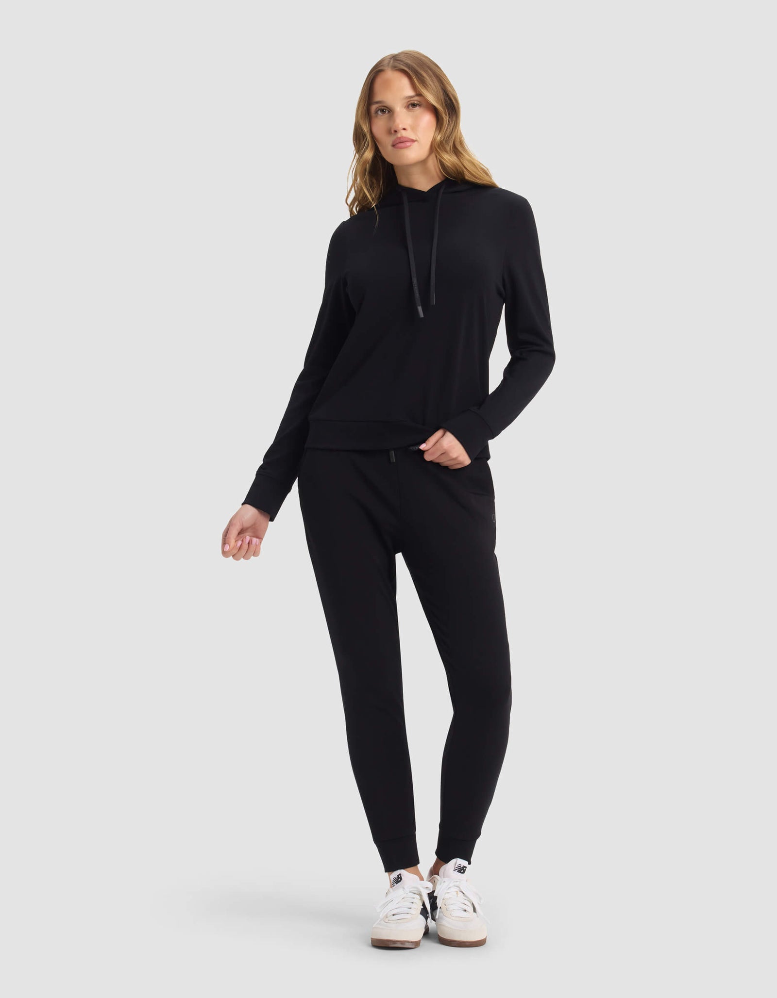 A woman stands against a light gray background wearing the Cozy Earth Women's Brushed Bamboo Hoodie & Jogger Set in black, paired with white sneakers. She poses with one hand in her pocket and a relaxed posture.