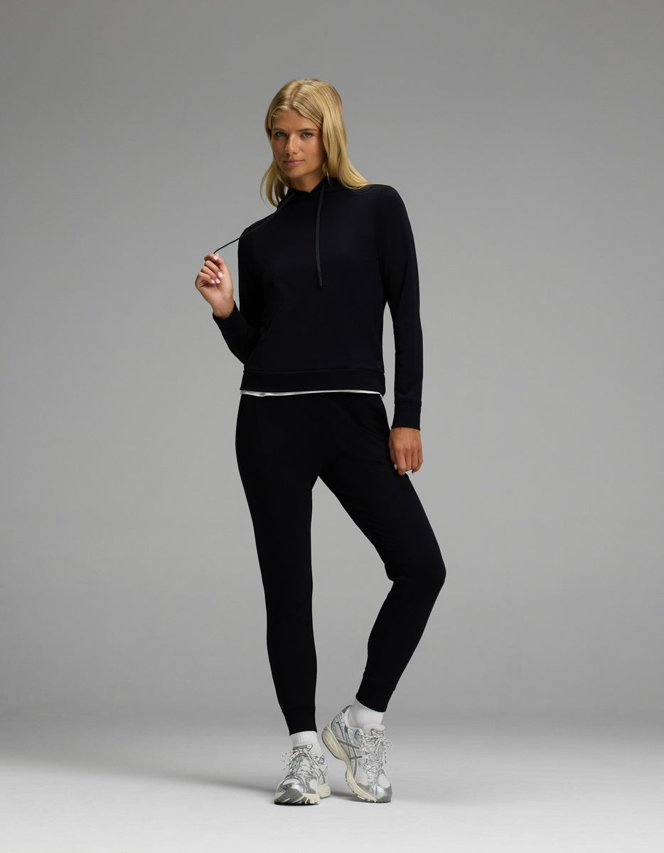 A woman stands confidently against a plain grey background wearing the Cozy Earth Women’s Brushed Bamboo Hoodie & Jogger Set in black and white sneakers, holding the hoodie’s drawstring.|Color:Black