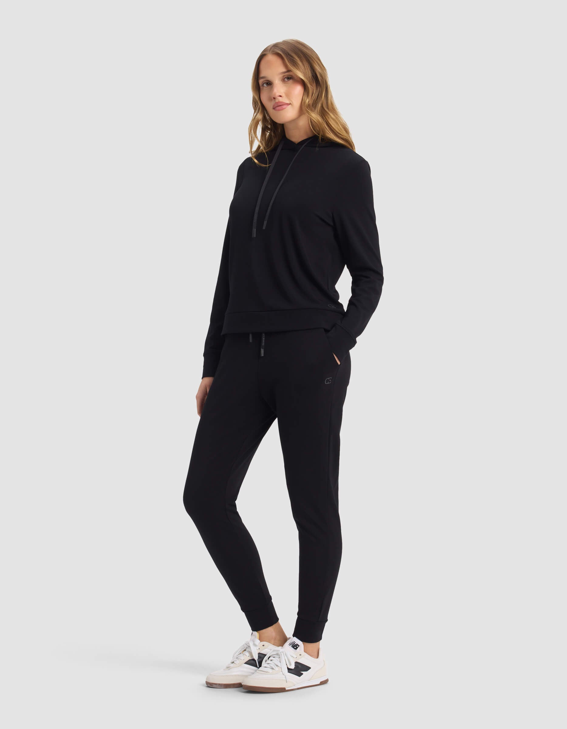 A woman faces forward against a light gray background, wearing Cozy Earth's Women's Brushed Bamboo Hoodie & Jogger Set in black, paired with white sneakers featuring black accents.