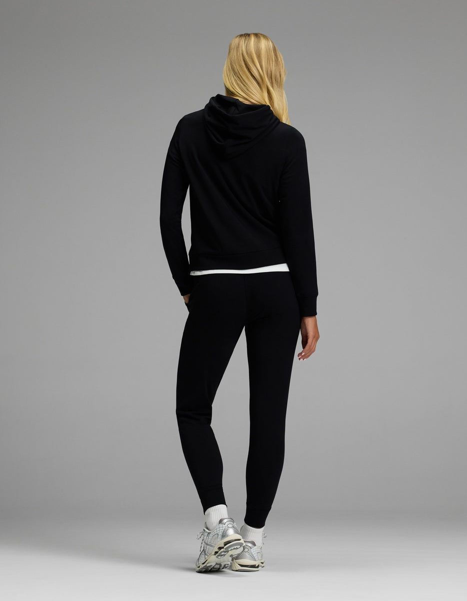 A person with long blonde hair, dressed in the Cozy Earth Women’s Brushed Bamboo Hoodie & Jogger Set in black and white sneakers, stands facing away against a plain gray background.