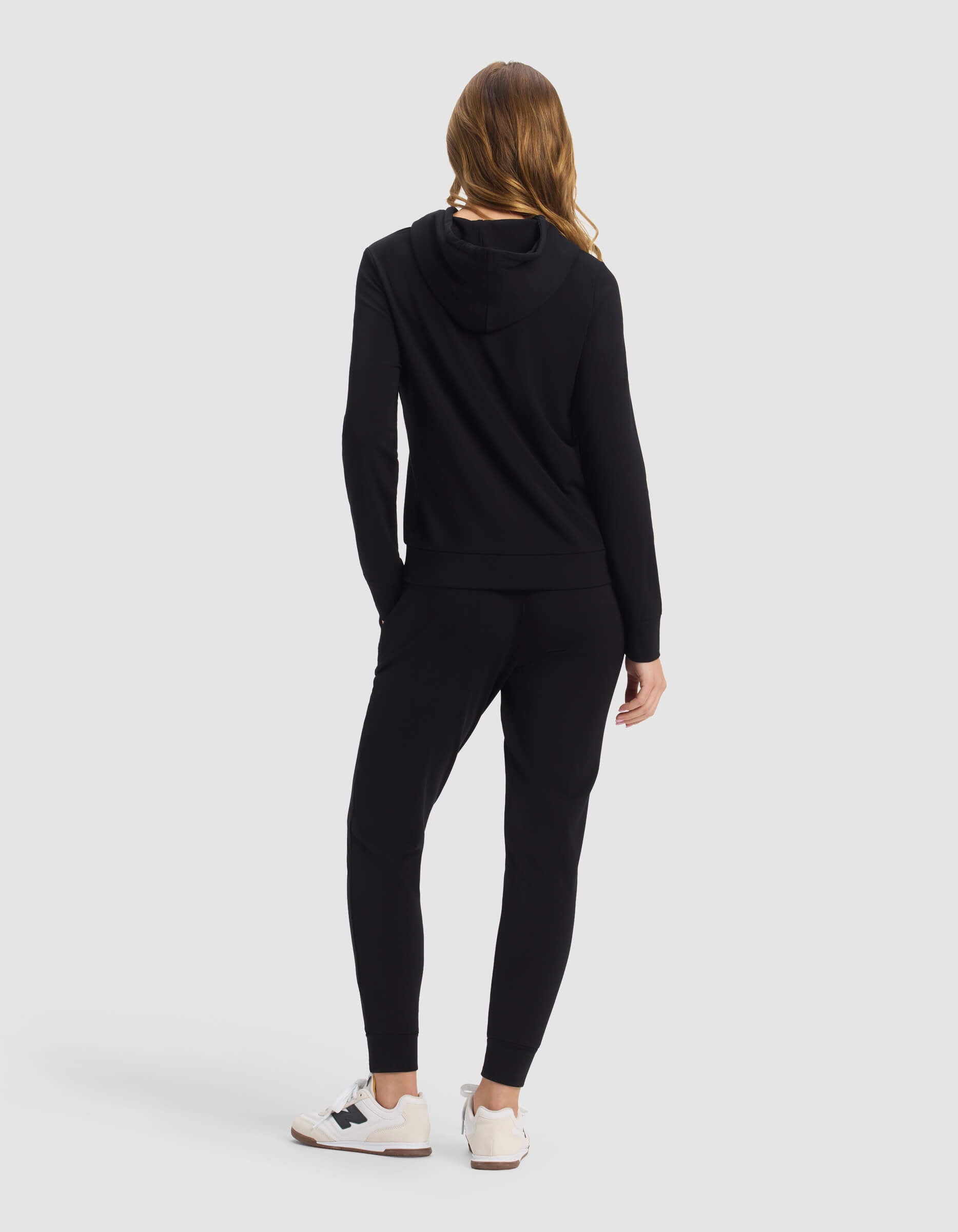 A woman with long hair is seen from behind wearing the Cozy Earth Women's Brushed Bamboo Hoodie & Jogger Set in black, paired with white sneakers, standing against a plain light gray background.