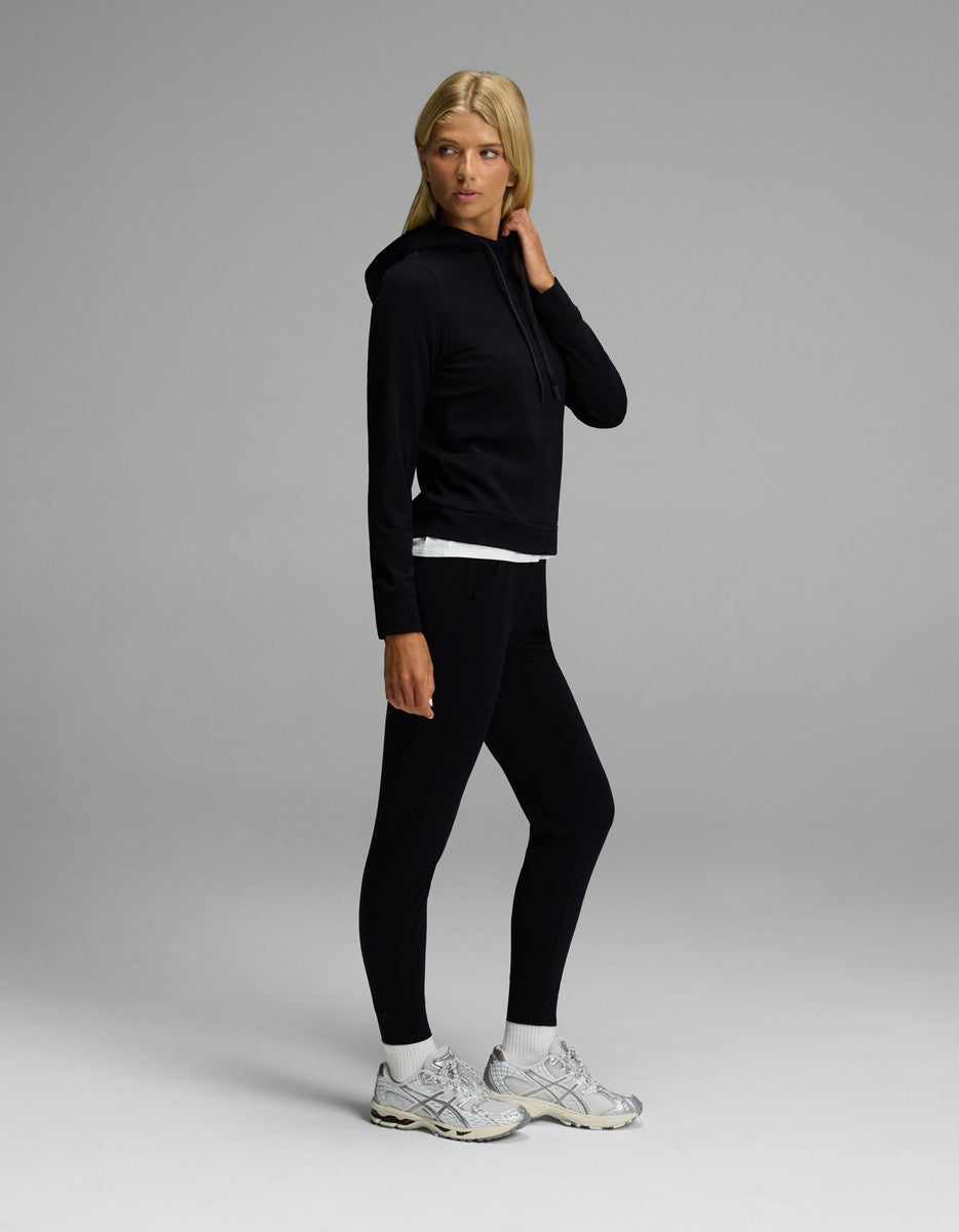 A person with long blonde hair wears the Cozy Earth Women’s Brushed Bamboo Hoodie & Jogger Set in black, paired with white sneakers and a white shirt peeking out, standing against a plain gray background.